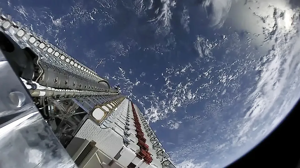 A long stack of flat Starlink satellites on a rocket's second stage, with the curve of the Earth and clouds behind it